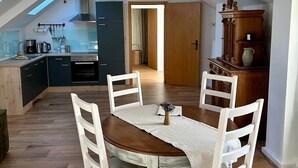 Een eetgedeelte met een houten tafel en witte stoelen, een keuken op de achtergrond en een schuin dak met dakramen.