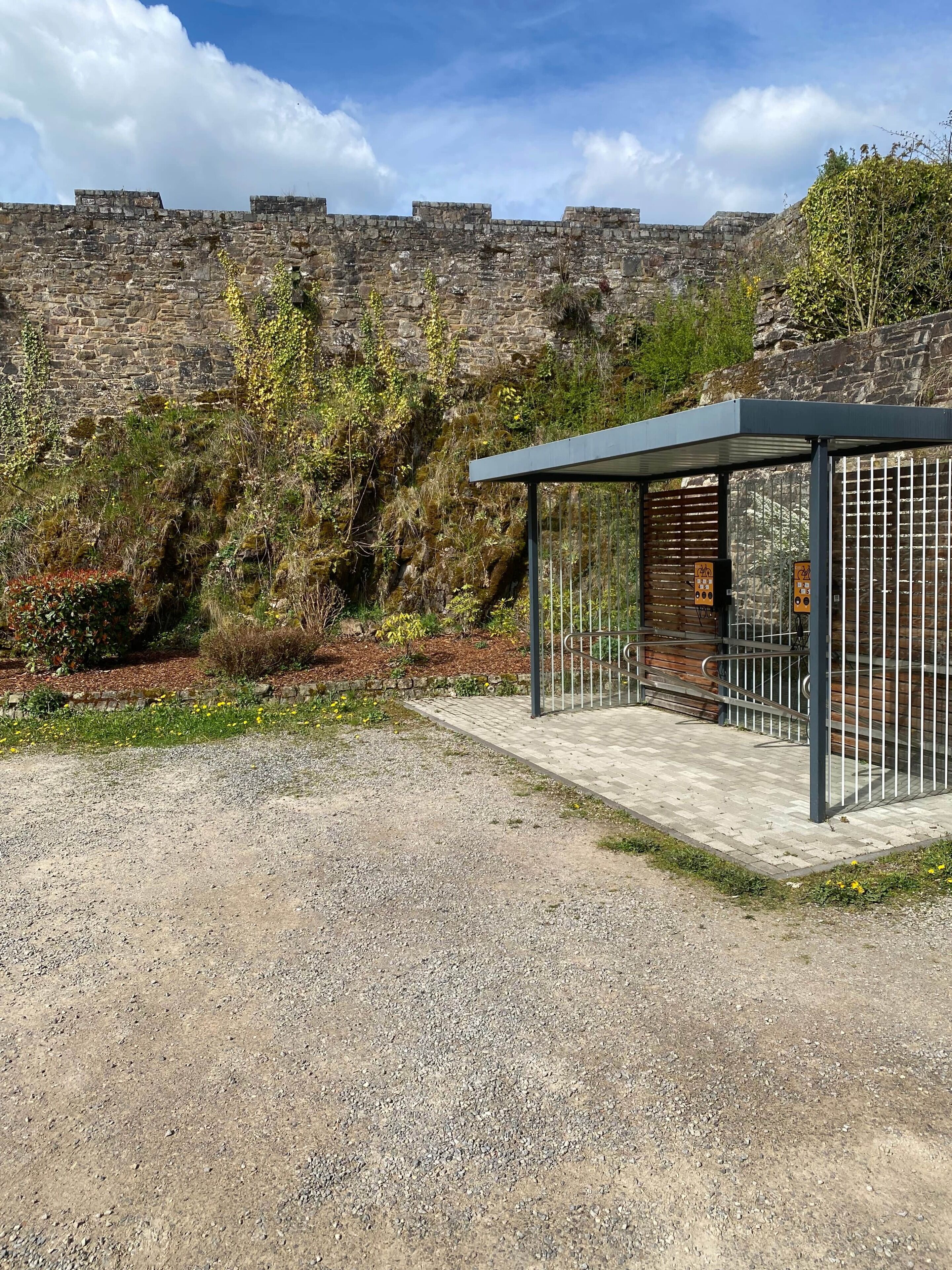 A covered outdoor seating area with a metal railing, a stone wall in the background, and a gravel ground.
