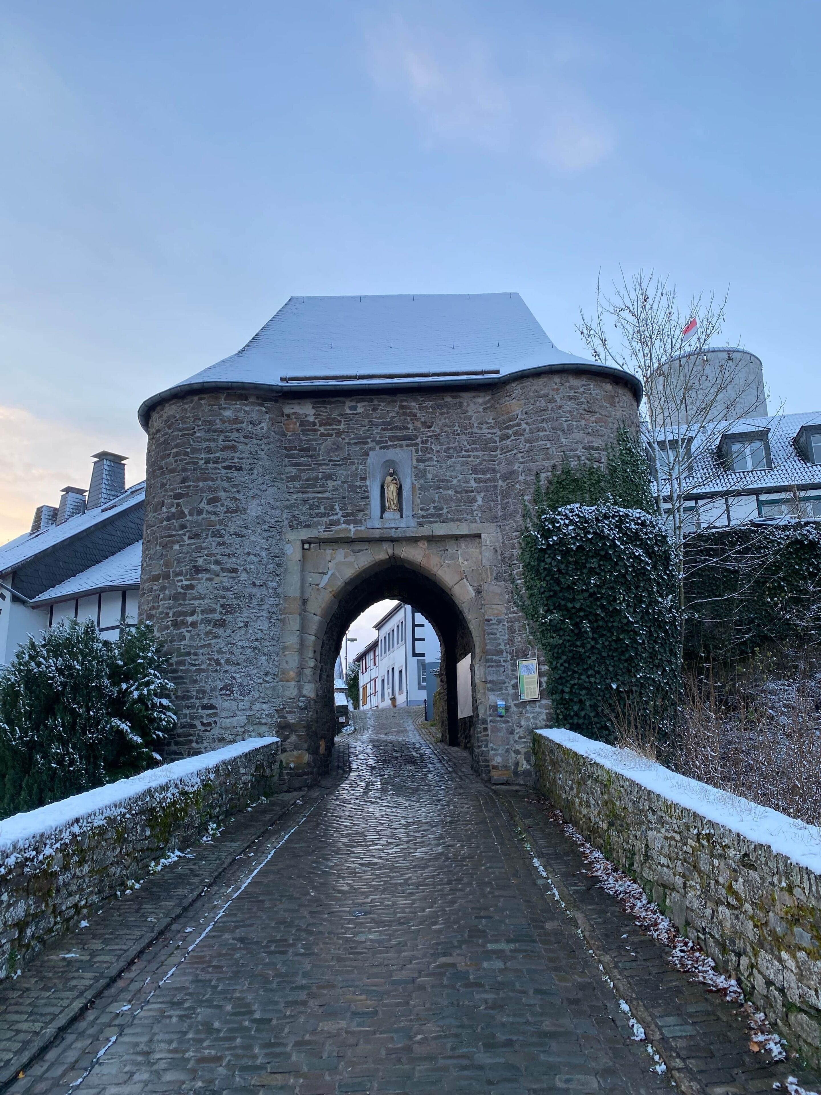Ein steinerner Bogen führt zu einer Kopfsteinpflasterstraße, umgeben von Gebäuden und schneebedeckten Bäumen.
