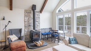 A cozy living room with a stone wall, a black wood-burning stove, a dining table with blue chairs, and a white sofa.