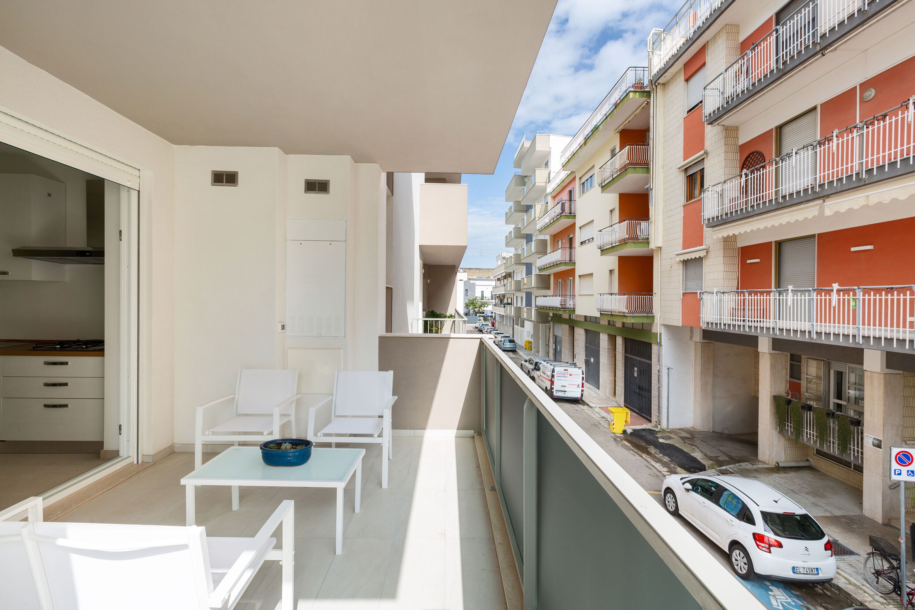 Ein Balkon mit weißer Möbel, einem Esstisch und Blick auf eine Straße mit parkenden Autos und Gebäuden.