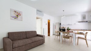 Un salon moderne avec un canapé marron, un coin repas avec une table et des chaises, et une cuisine avec des meubles blancs et un réfrigérateur en acier inoxydable.