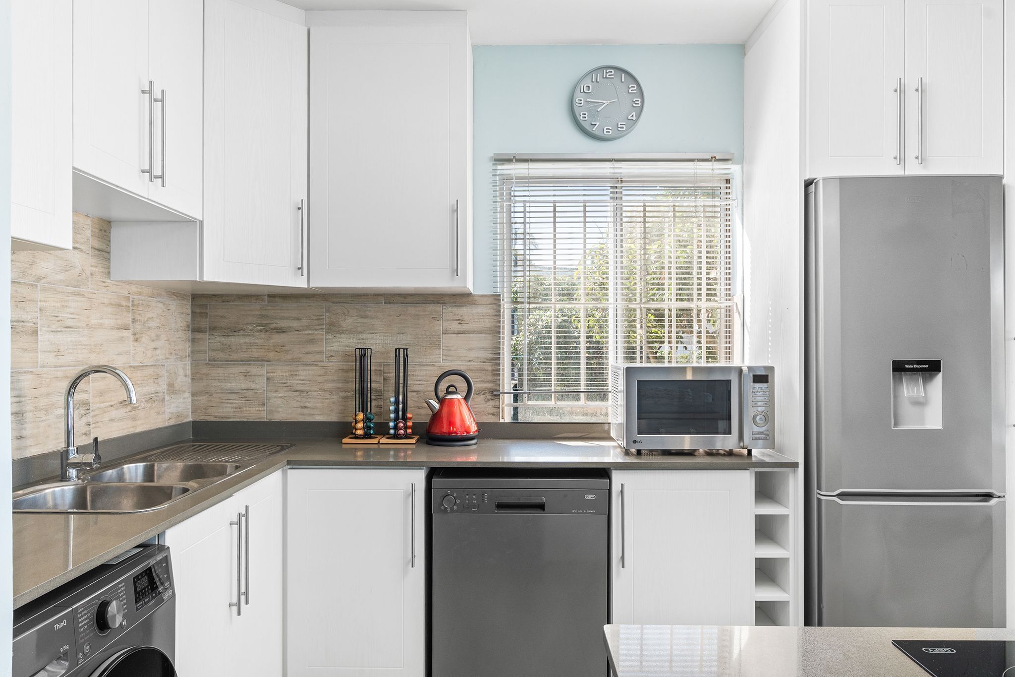 Una cocina moderna con armarios blancos, un refrigerador de acero inoxidable, un lavavajillas, un microondas y un hervidor.
