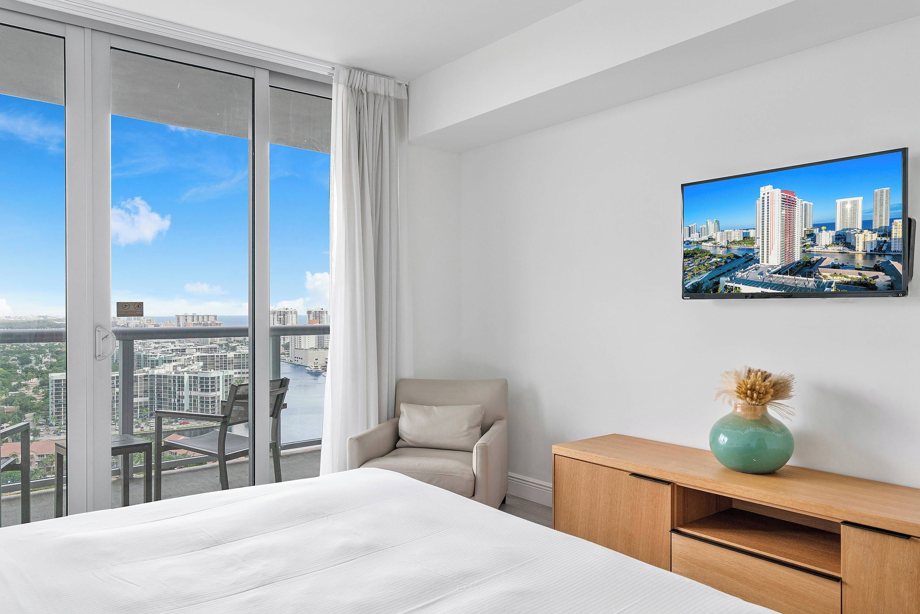 A hotel room with a large bed, a chair, a wooden side table, a vase, and a cityscape painting on the wall.