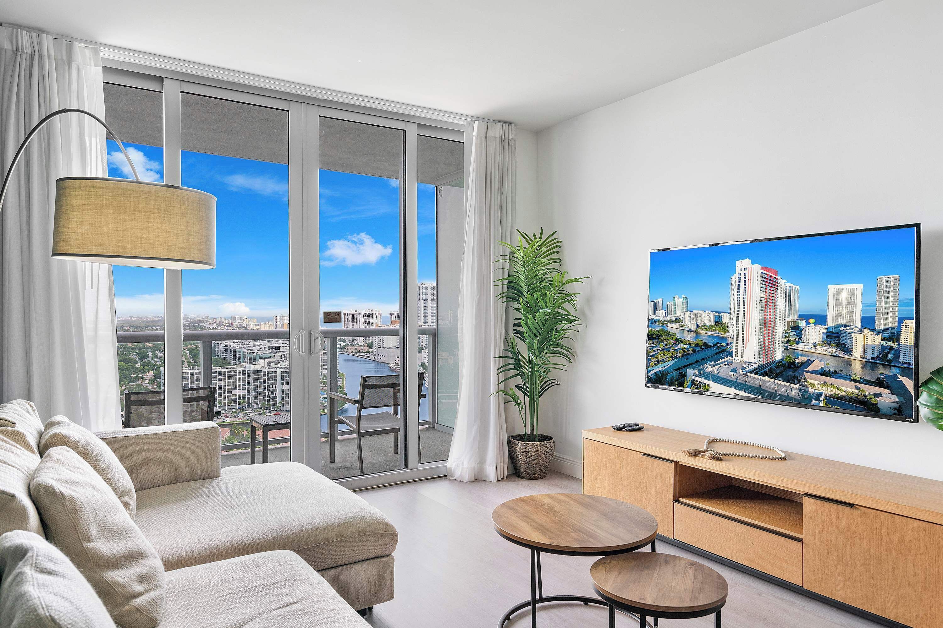 Un salon moderne avec un canapé, un téléviseur, une plante et une vue sur le paysage urbain, visible à travers de grandes fenêtres.