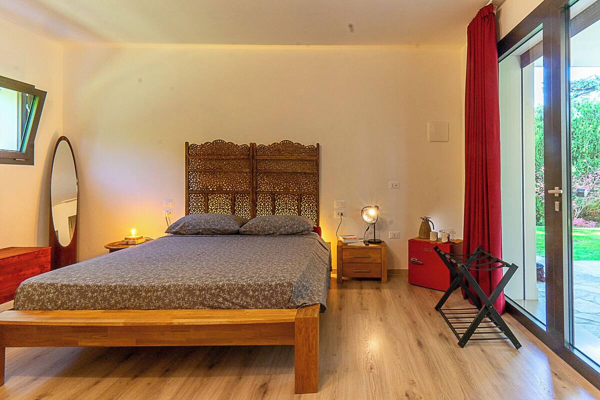 A bedroom with a wooden bed, a nightstand with a lamp, a red cabinet, and a sliding glass door to a garden.
