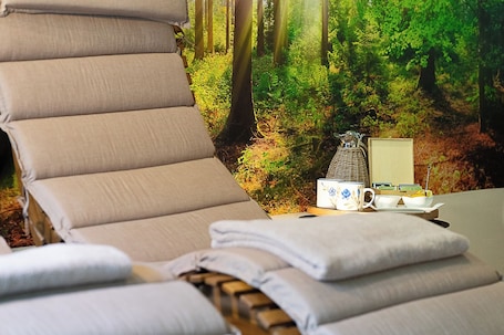 A sun lounger with a forest mural backdrop, a small table with a pitcher, a mug, and a book.. Marimastè