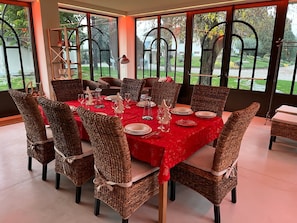 Un coin repas avec une nappe rouge, des chaises en osier et une vue sur un jardin grĂące Ă de grandes fenĂȘtres.