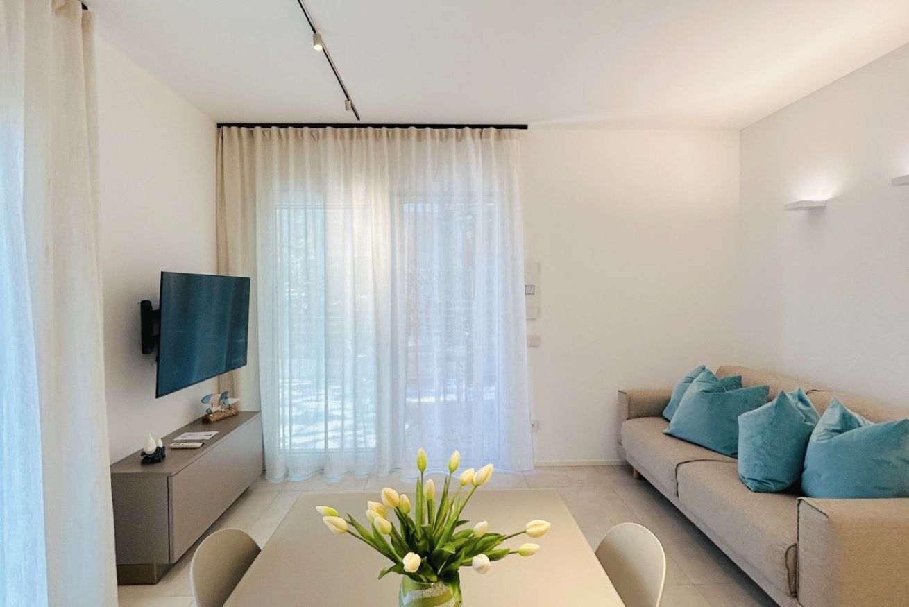 Ein modernes Wohnzimmer mit Sofa, Fernseher, Esstisch mit Blumenvase und einem großen Fenster mit durchsichtigen Vorhängen.