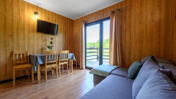 A room with wooden walls, a flat-screen TV mounted on the wall, a dining table with chairs, and a sofa with cushions.