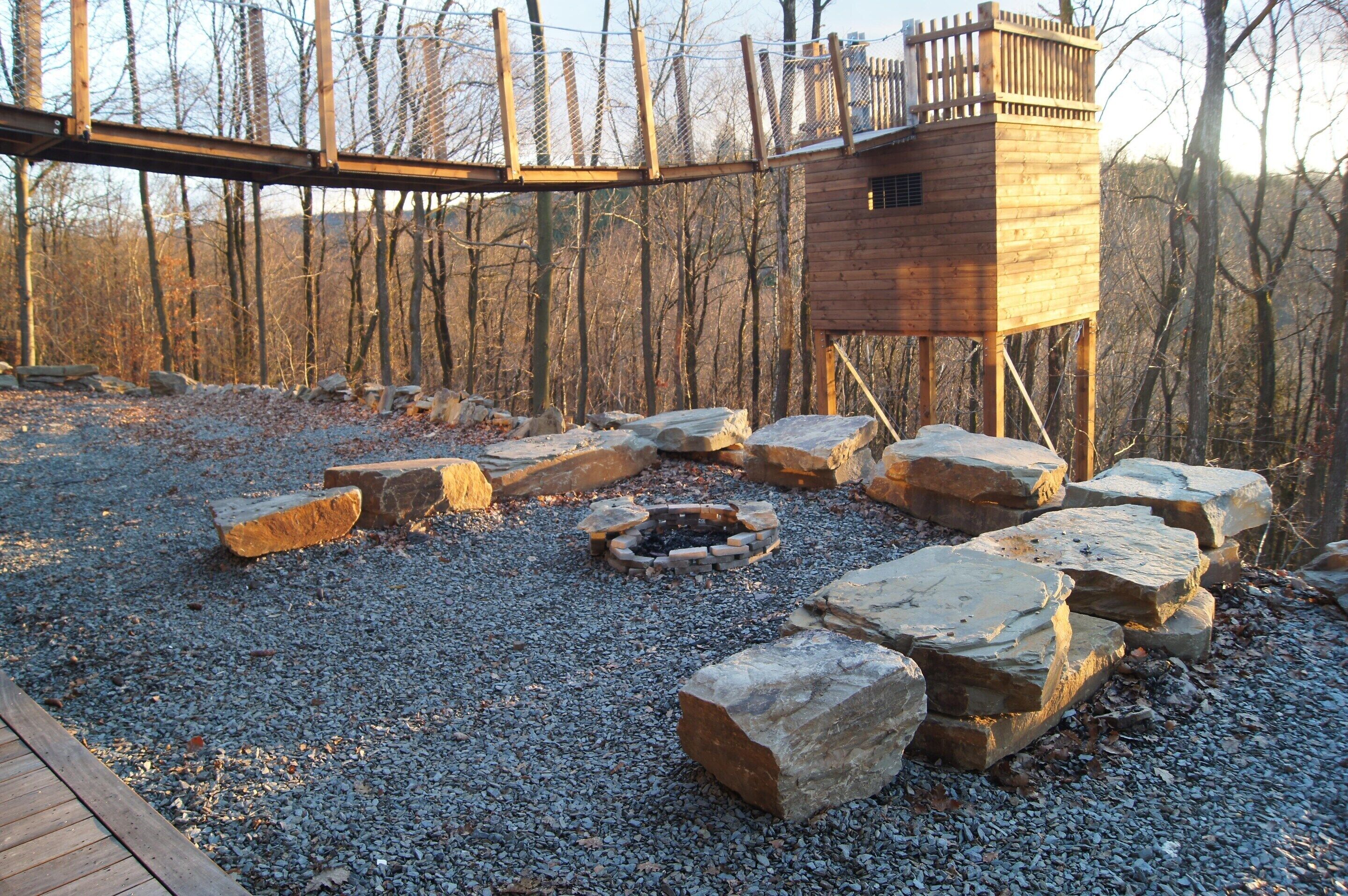 A wooden cabin with a stone fire pit surrounded by rocks, set in a forested area with a wooden deck.
