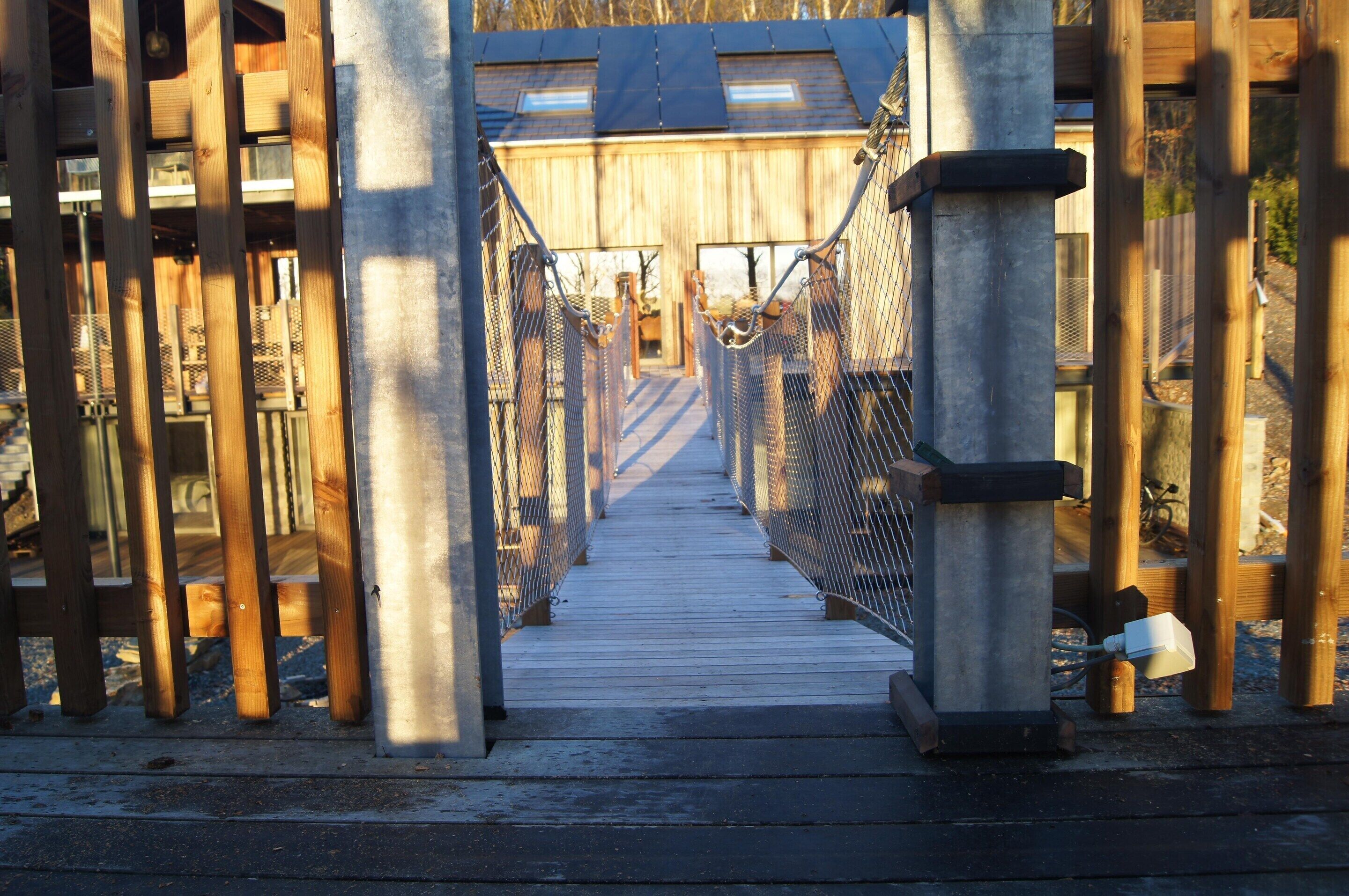 Un pont suspendu en bois, doté de garde-corps métalliques et d’un pilier en béton.