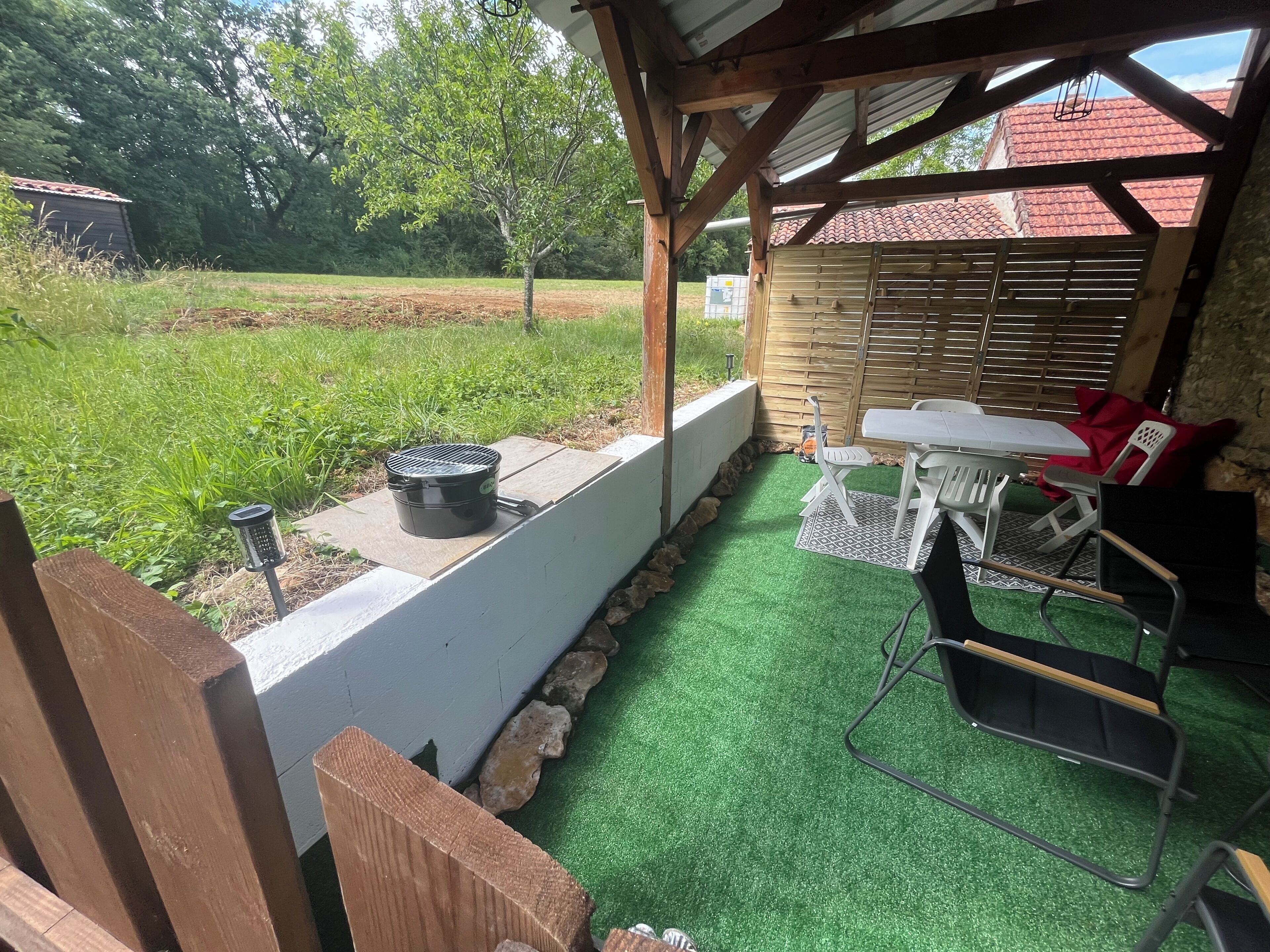 A covered outdoor patio with a table and chairs, a grill, and a garden view.