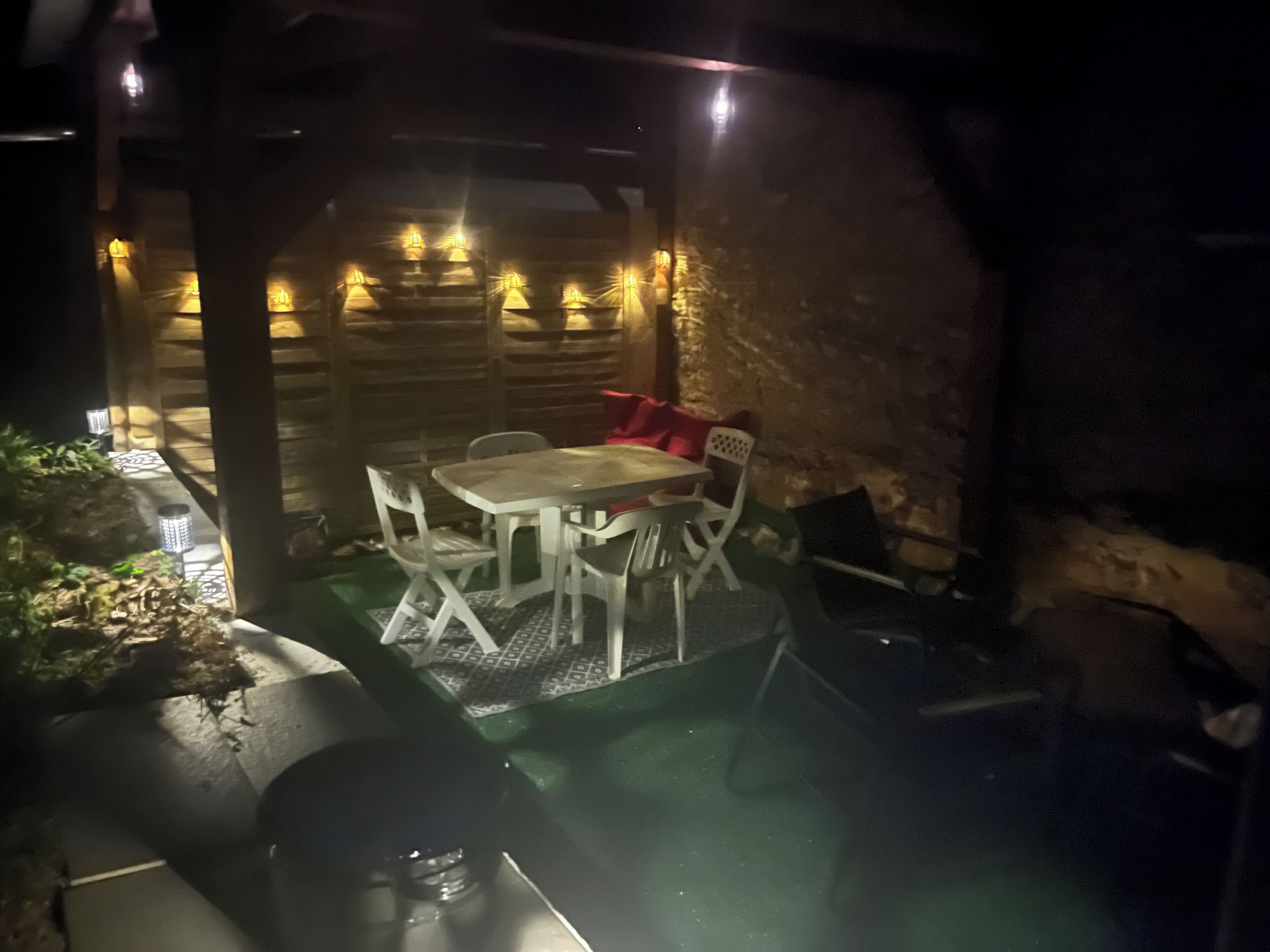 An outdoor patio area with a table and chairs, surrounded by stone walls and illuminated by string lights.