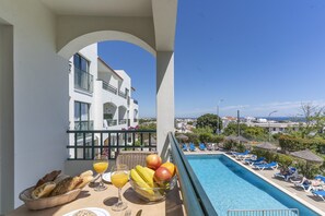 A balcony with a table set for breakfast, a swimming pool, and a view of a white building with balconies. - #384 Bright Pool View Apartment | AC & Balcony (Albufeira)