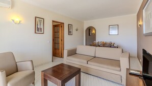 Ein Wohnzimmer mit einer Couch, einem Couchtisch und einem Fernseher. An den Wänden befinden sich Bilder in Rahmen und eine Tür mit Glasfeldern.