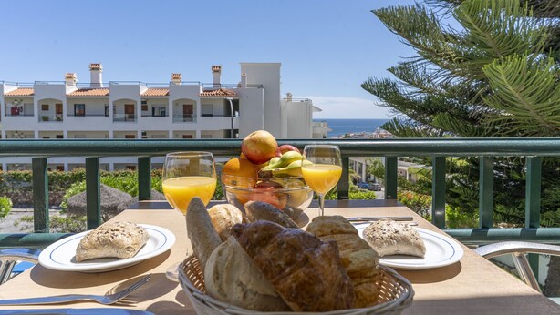Ein Esstisch mit Brot, Obst und Orangensaft, mit Blick auf ein Küstengebäude und Palmen.
