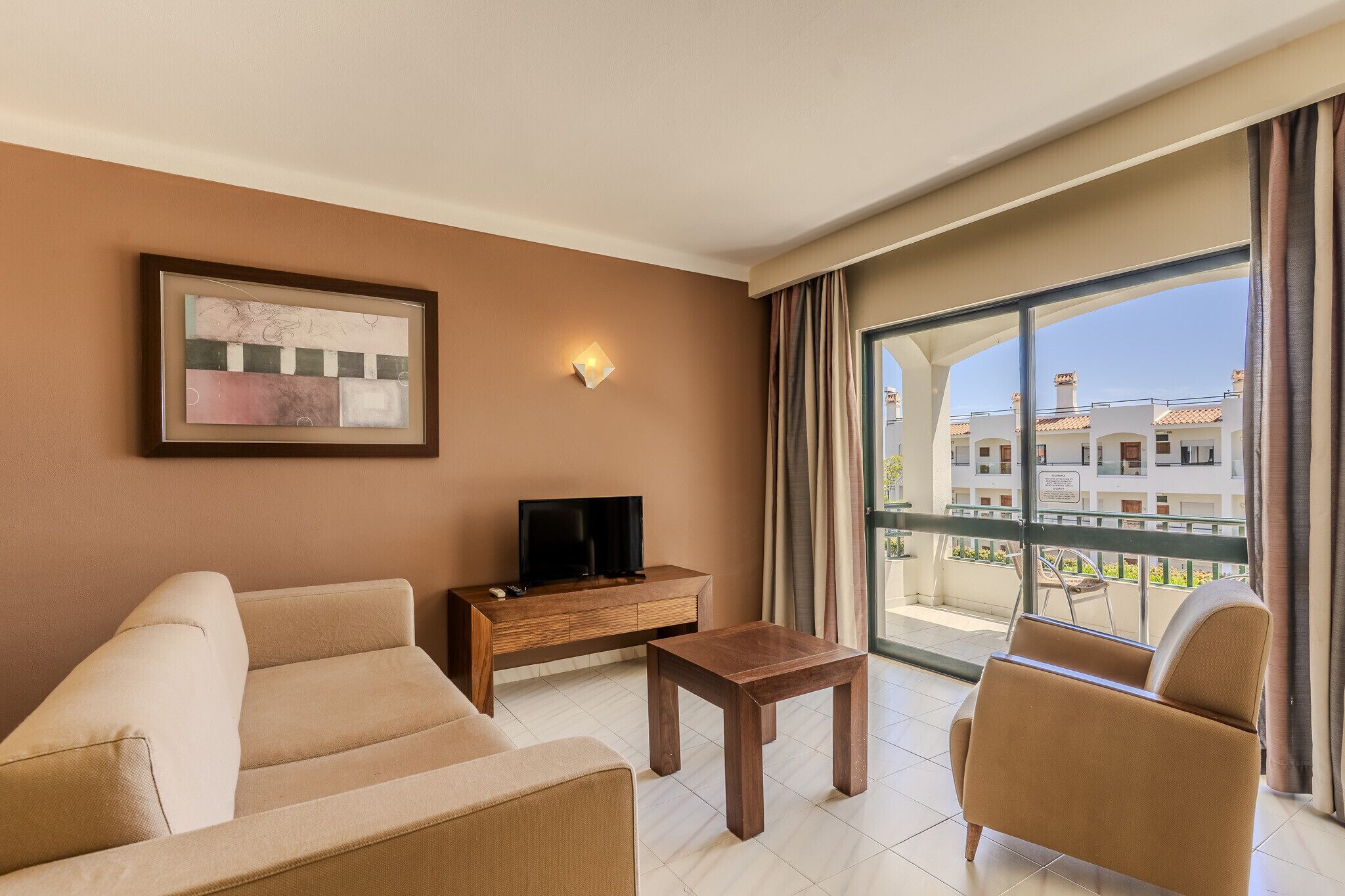 Une chambre d’hôtel avec un canapé, une petite table en bois, un téléviseur posé sur un meuble en bois et un balcon donnant sur des immeubles.
