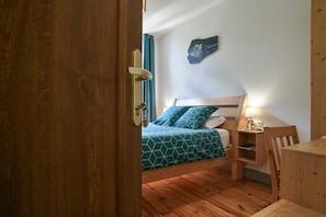 A room with a wooden floor, a bed with a geometric-patterned bedspread, a bedside table with a lamp, and a wall-mounted artwork.