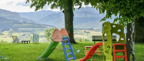 A playground with slides and climbing structures, surrounded by a grassy field and a tree, with mountains in the background.