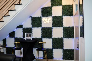 Un escalier menant à une pièce dont le mur est décoré de motifs à carreaux, orné d'un néon et comprenant une table avec des tabourets.