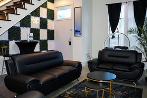 Un salon moderne avec un canapé en cuir noir, une table basse ronde et un fauteuil noir. On y trouve un escalier avec des rampes noires et un néon mural.