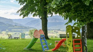 Un parque infantil con toboganes y estructuras para escalar, rodeado de un césped y un árbol, con montañas al fondo.