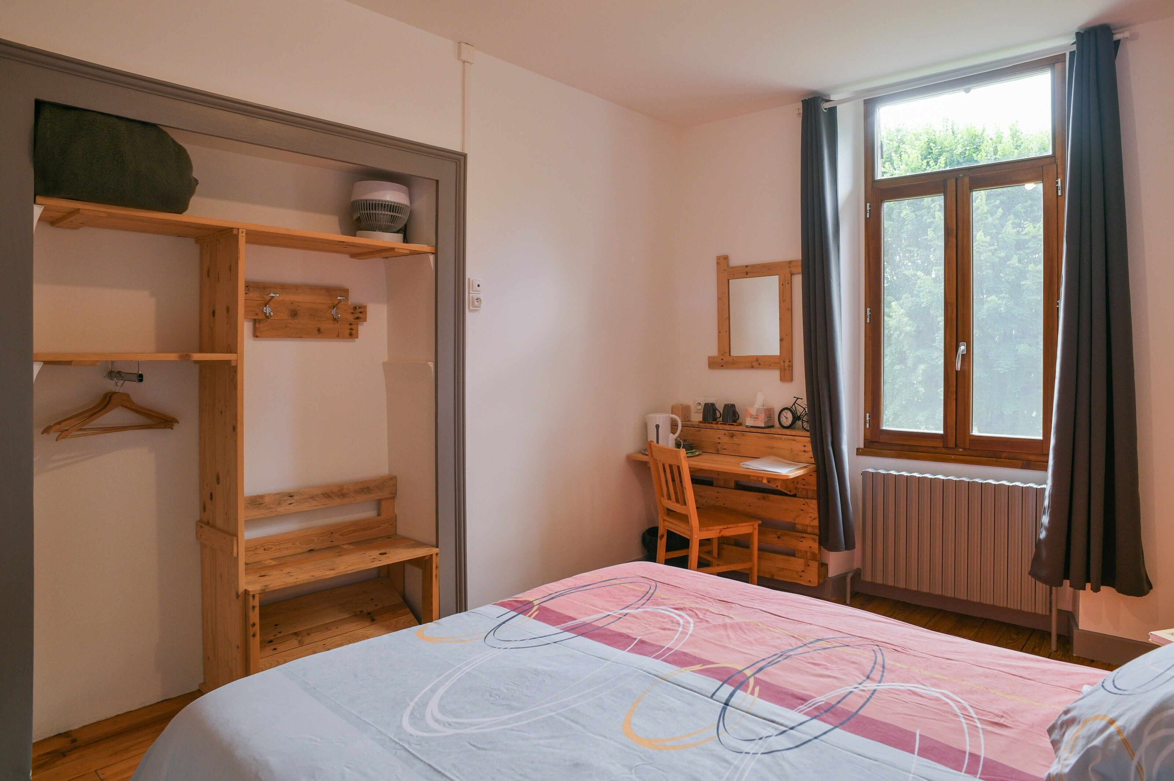 Une chambre à coucher comprenant un lit, un bureau en bois, une chaise, un radiateur, une fenêtre avec des rideaux et une armoire avec des étagères.