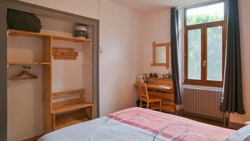 Une chambre à coucher comprenant un lit, un bureau en bois, une chaise, un radiateur, une fenêtre avec des rideaux et une armoire avec des étagères.