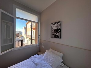 Une chambre Ă coucher avec un lit, une fenĂȘtre donnant sur des immeubles et un tableau encadrĂ© accrochĂ© au mur.