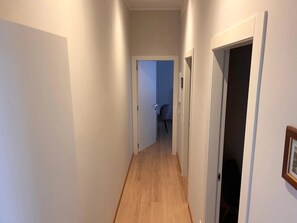 Um corredor estreito com piso de madeira e paredes brancas, que leva a um quarto com uma porta à direita.