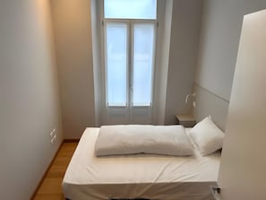 Una camera da letto piccola e pulita con un letto, un comodino, una finestra con tende e una lampada a parete.