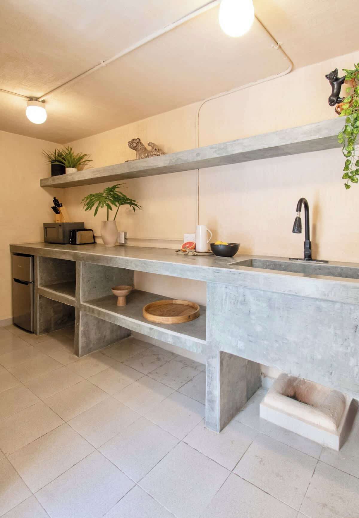 A kitchen with a concrete countertop, a built-in sink, a refrigerator, and a shelf with plants.