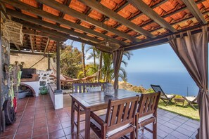 A covered outdoor seating area with a table and chairs, a grill, and a view of the ocean.