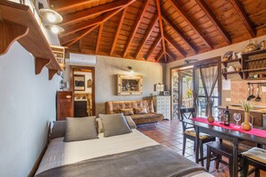 A bedroom with a large bed, a wooden ceiling, a dining area with a table and chairs, and a kitchen area with shelves and a counter.