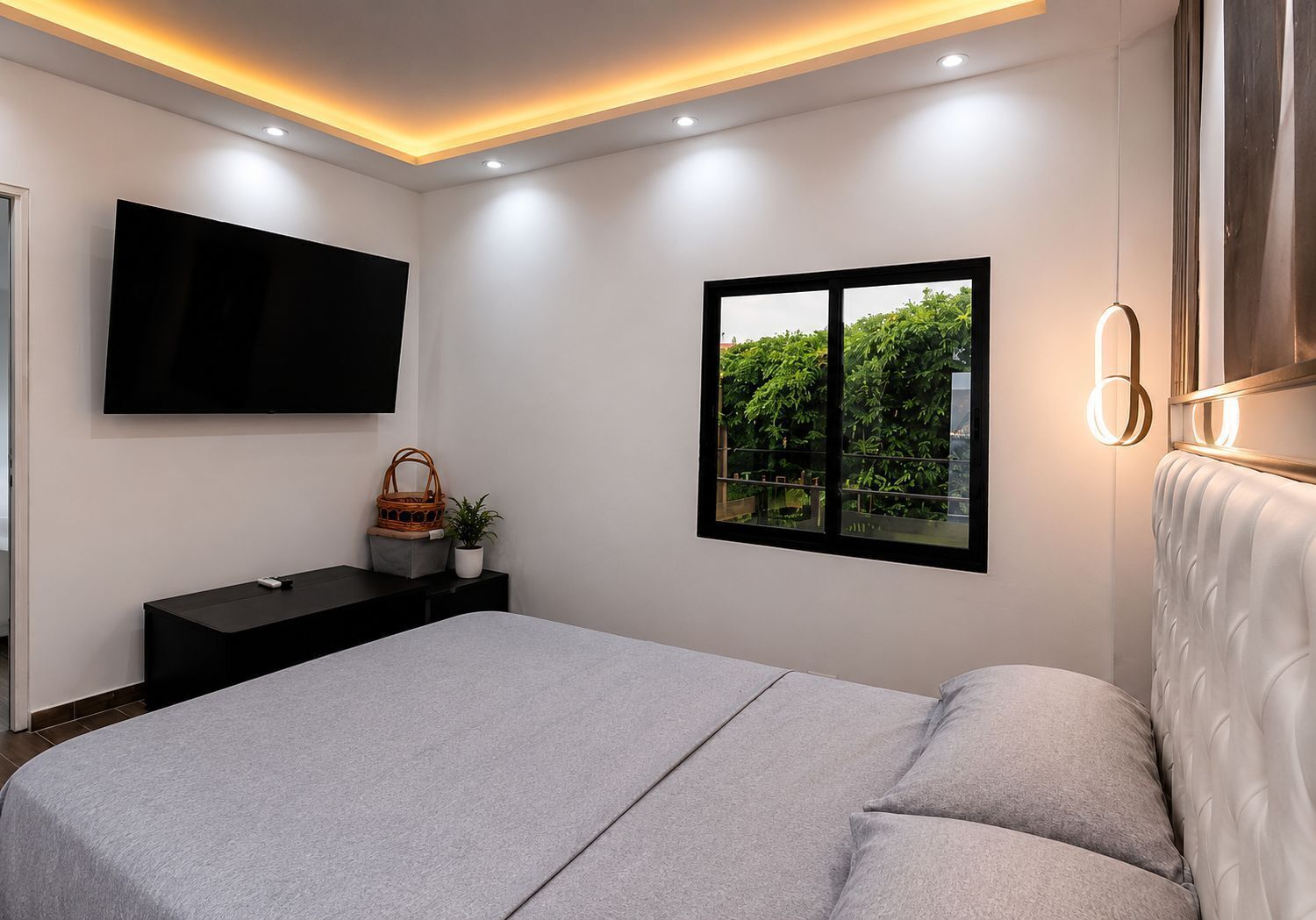 A modern bedroom with a bed, a TV mounted on the wall, a window with a view of greenery, and a bedside table with a plant.