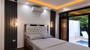 A modern bedroom with a tufted headboard, a bed with grey bedding, a built-in air conditioning unit, and a glass door leading to a pool area.