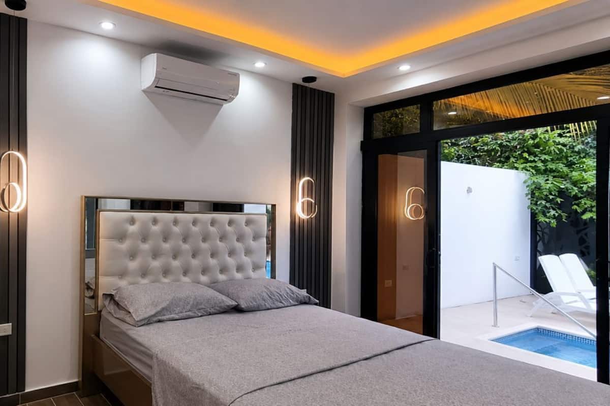 Une chambre moderne avec une tête de lit capitonnée, un lit avec une literie grise, un système de climatisation intégré et une porte en verre donnant sur un espace piscine.