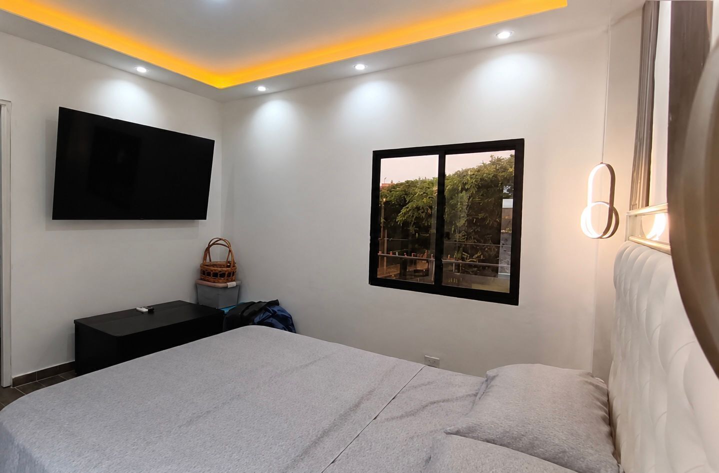 A modern bedroom with a bed, a TV mounted on the wall, a window with a view, and a bedside table with a basket.