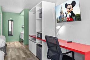 Una habitación de hotel con un escritorio rojo, una silla de oficina negra, un televisor mostrando a Mickey Mouse y un espejo.