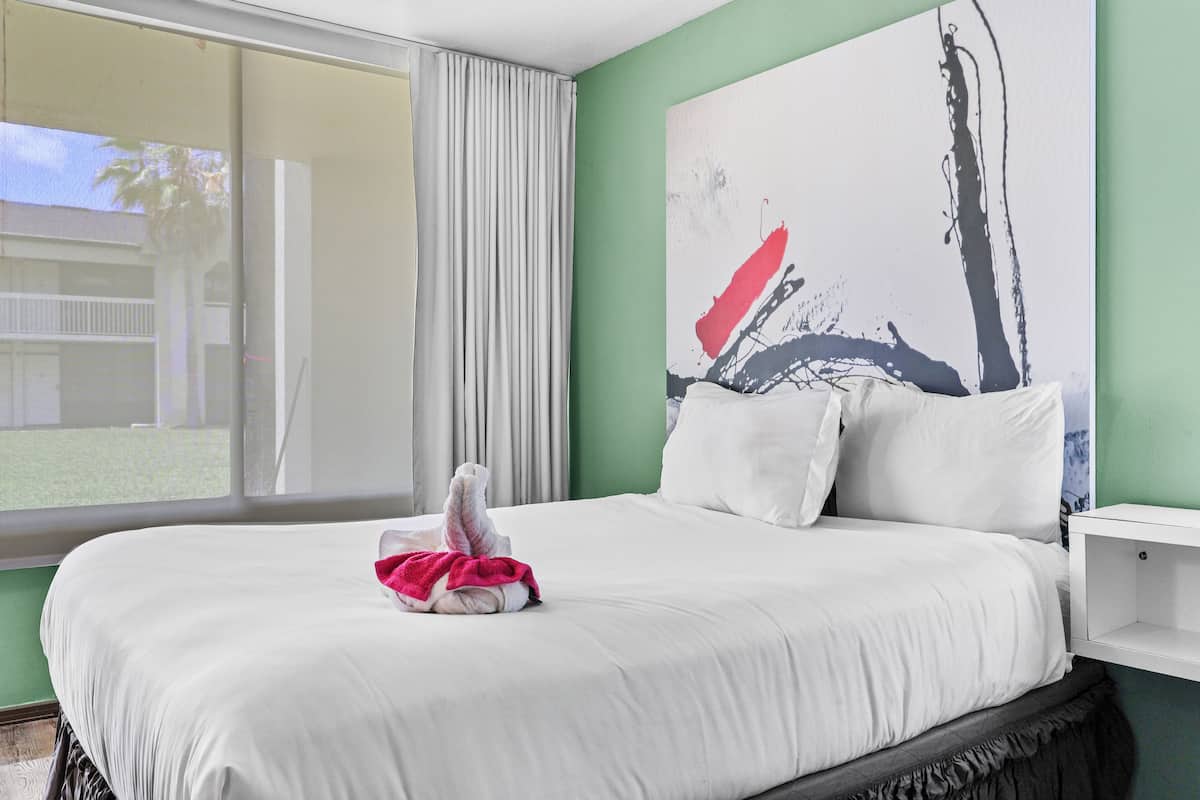 A hotel room with a large bed, a window with curtains, a green wall, and a piece of abstract art.