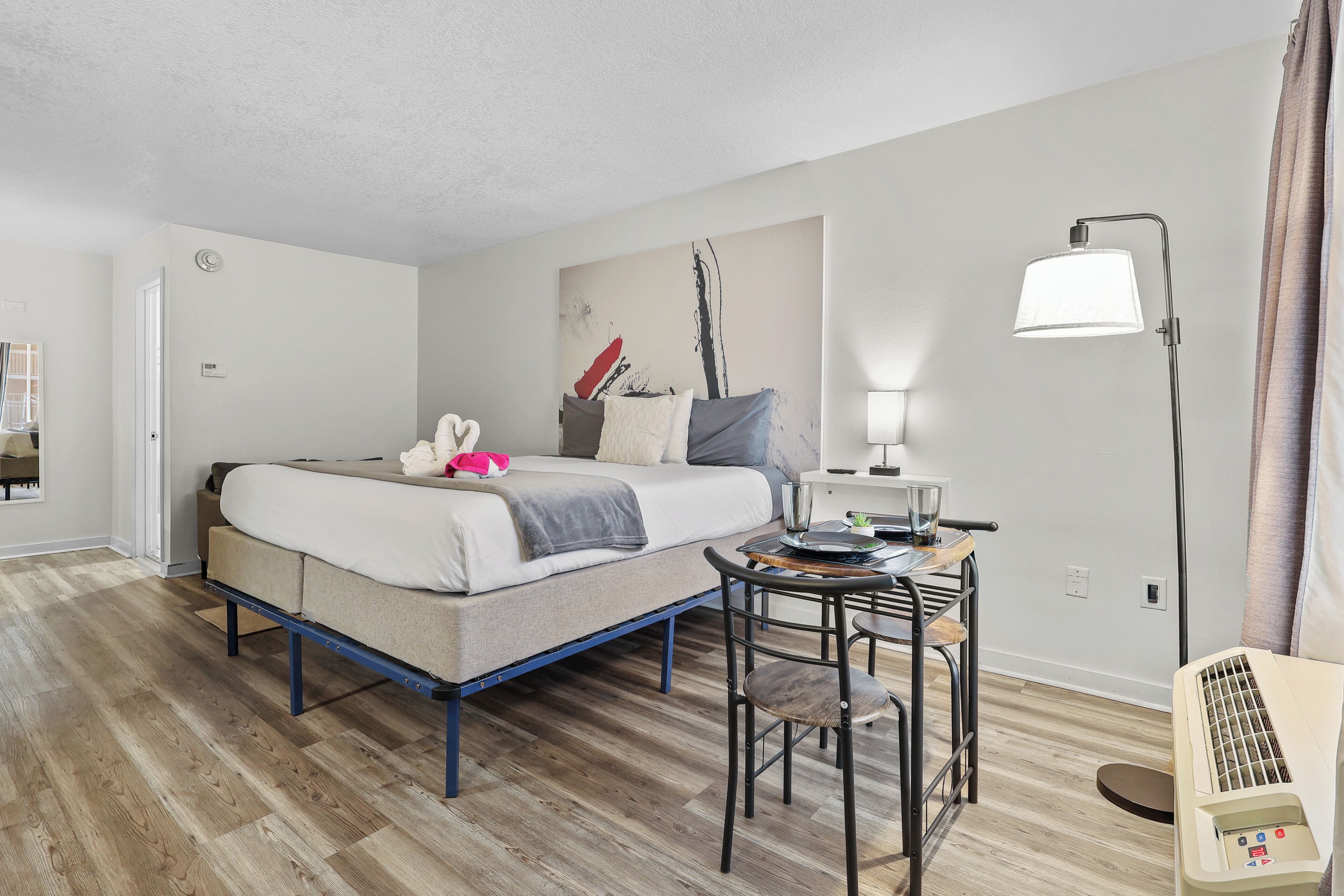 A hotel room with a bed, a desk with a lamp, a chair, and a heater.