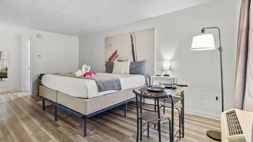 A hotel room with a bed, a desk with a lamp, a chair, and a heater.