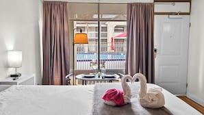 Hotellihuone, jossa on sänky, lamppuvarustettu pöytä ja ikkunasta avautuva näkymä uima-altaalle ja ympäröiville rakennuksille.