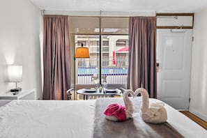 Habitación de hotel con cama, una mesa con lámpara, una toalla con forma de cisne y vistas a una piscina y un edificio a través de una puerta de cristal.