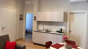 A compact kitchen with a sink, cabinets, and an air conditioner. There is a dining area with a table and chairs, and a sofa with a red cushion. - Apartment 'Verdi' with Wi-Fi and Air Conditioning (Gorizia)