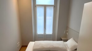 Een kleine, schone slaapkamer met een bed, een nachtkastje, een raam met gordijnen en een wandlamp.