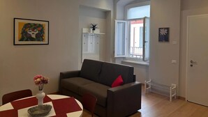 Een woonkamer met een grijze bank, een eettafel met een rood-witte tafelkleed, een vaas met bloemen en een raam met een open deur.