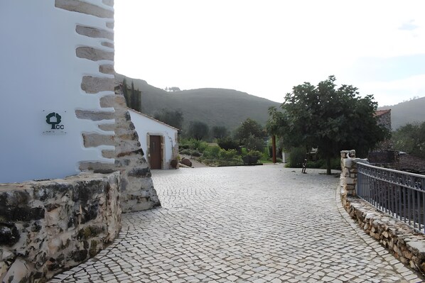 A cobblestone pathway leading to a white building with a door, surrounded by greenery and a stone wall.