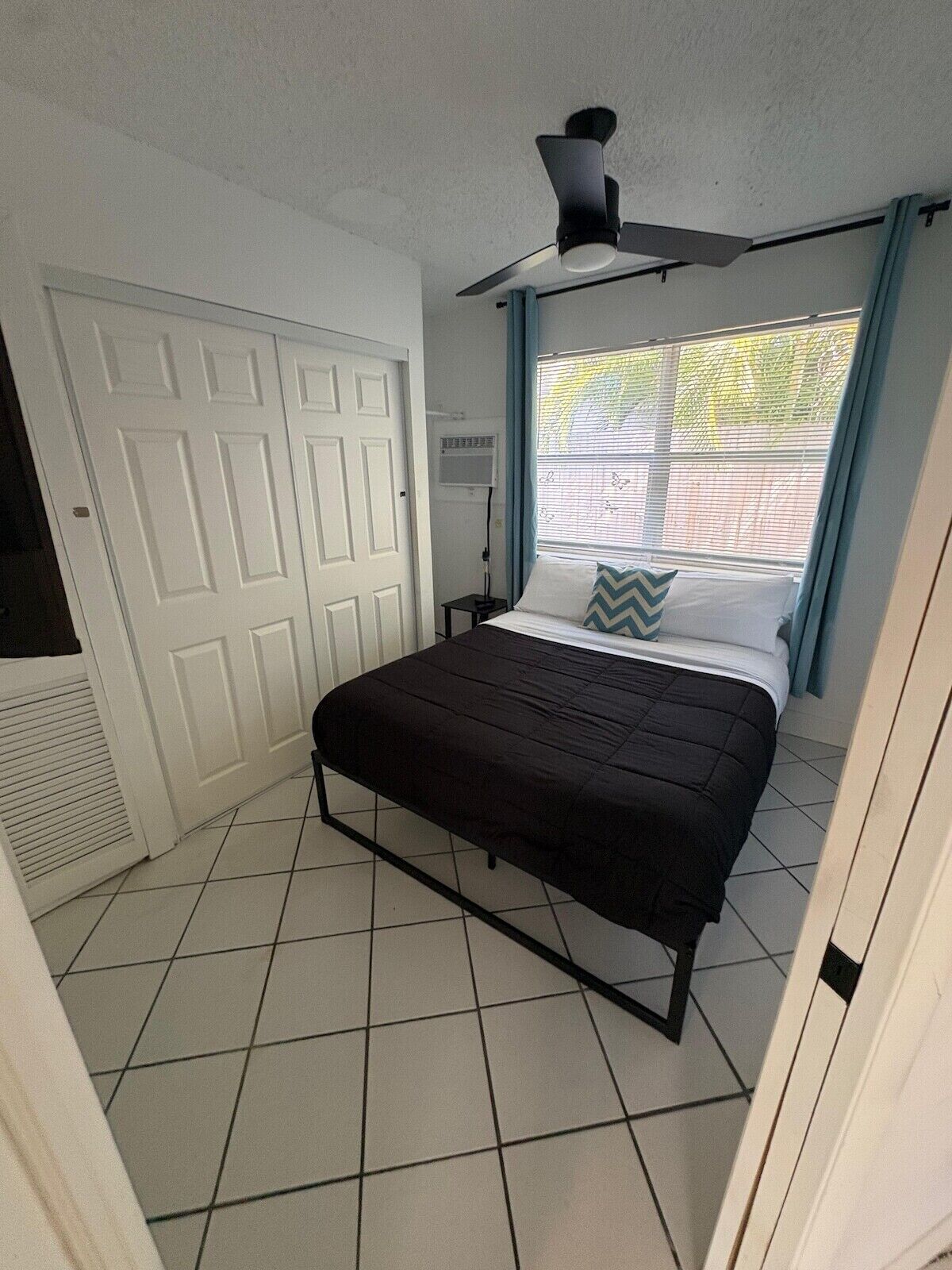 Une chambre à coucher avec un lit, un ventilateur de plafond, une fenêtre avec des stores et une porte blanche.